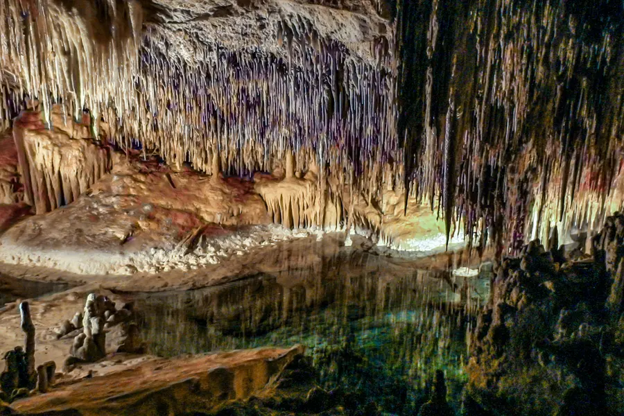 Weite Halle der Coves del Drac mit dicht hängenden Stalaktiten über einem spiegelnden unterirdischen See, warm angestrahlt.
