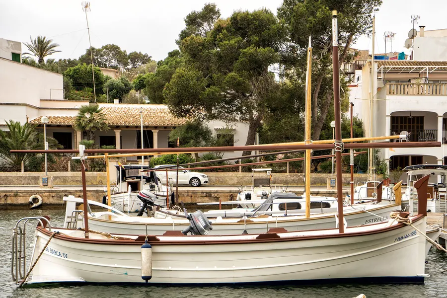 Ein hölzernes Boot mit Mast auf Böcken an Land in Porto Petro, dahinter weitere Boote und die Bäume am Ufer.