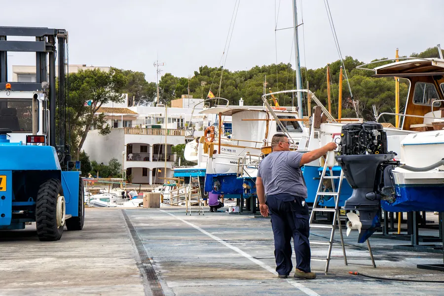 Ein Mann arbeitet auf einer Leiter an einem aufgebockten Boot am Kai von Porto Petro, daneben Werkzeug und ein Lieferwagen.
