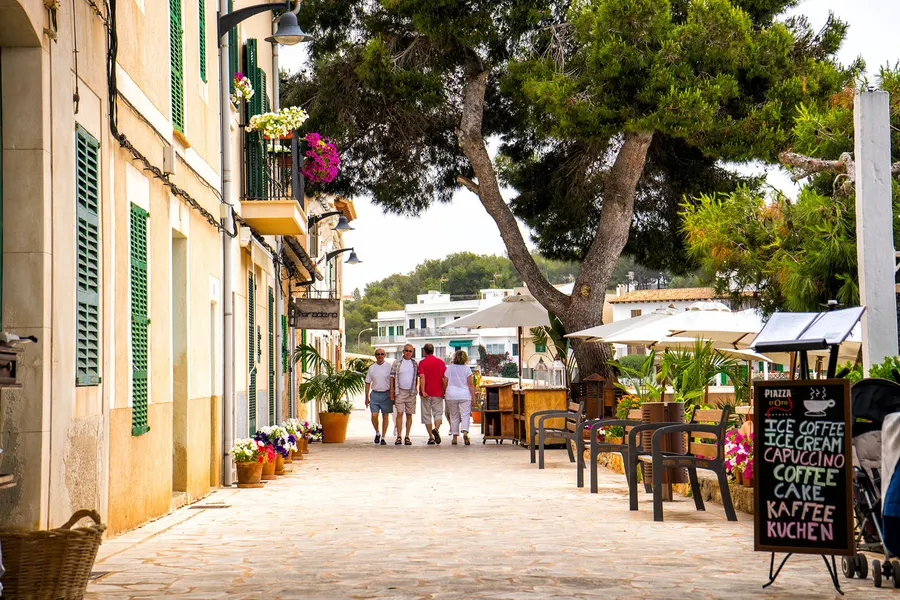 Restaurantterrasse unter hohen Bäumen an der Uferstraße von Porto Petro mit Tischen, Speisekarte und Gästen im Schatten.
