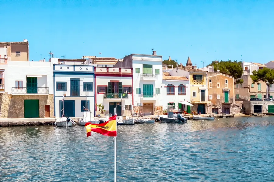 Bunte Häuserzeile mit gelben, grünen und blauen Fassaden direkt am Wasser von Portocolom, davor kleine Boote und eine spanische Flagge.
