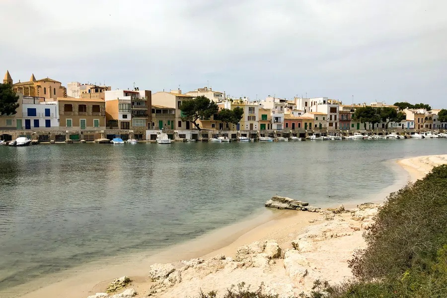 Weite Hafenbucht von Portocolom mit ruhigem Wasser, im Hintergrund die niedrige, bunte Uferzeile des Orts, im Vordergrund ein kleiner Strand.