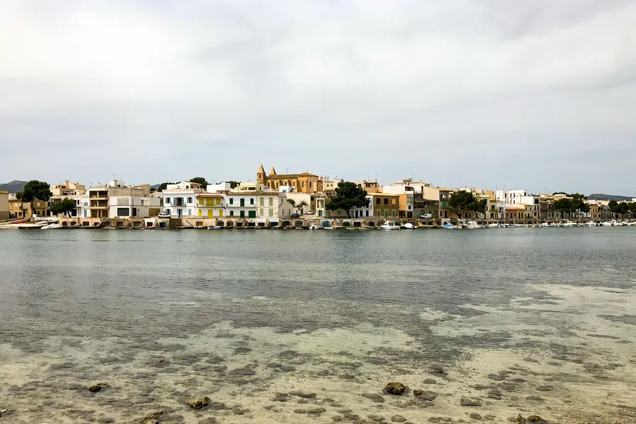 Blick über klares Flachwasser mit Steinen auf dem Grund hinüber zur Häuserzeile von Portocolom mit Bootshäusern und Kirchturm.