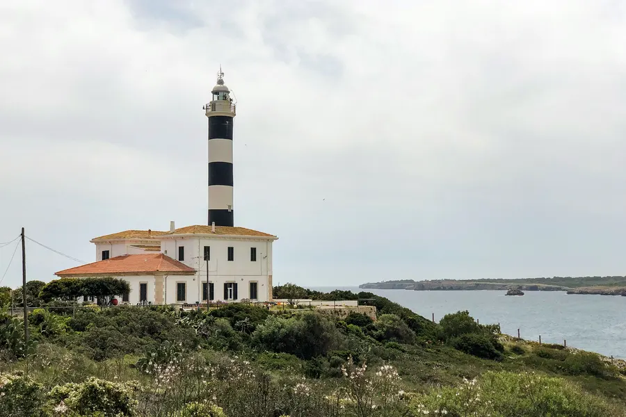 Der schwarz-weiß geringelte Leuchtturm von Portocolom mit dem weißen Wärterhaus auf der bewachsenen Landzunge, dahinter das offene Meer.