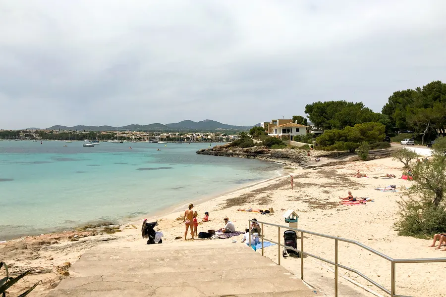 Badestelle in der Bucht von Portocolom mit Badegästen auf dem Sand, Geländer und Treppe zum Wasser, im Hintergrund Boote und die Uferzeile.