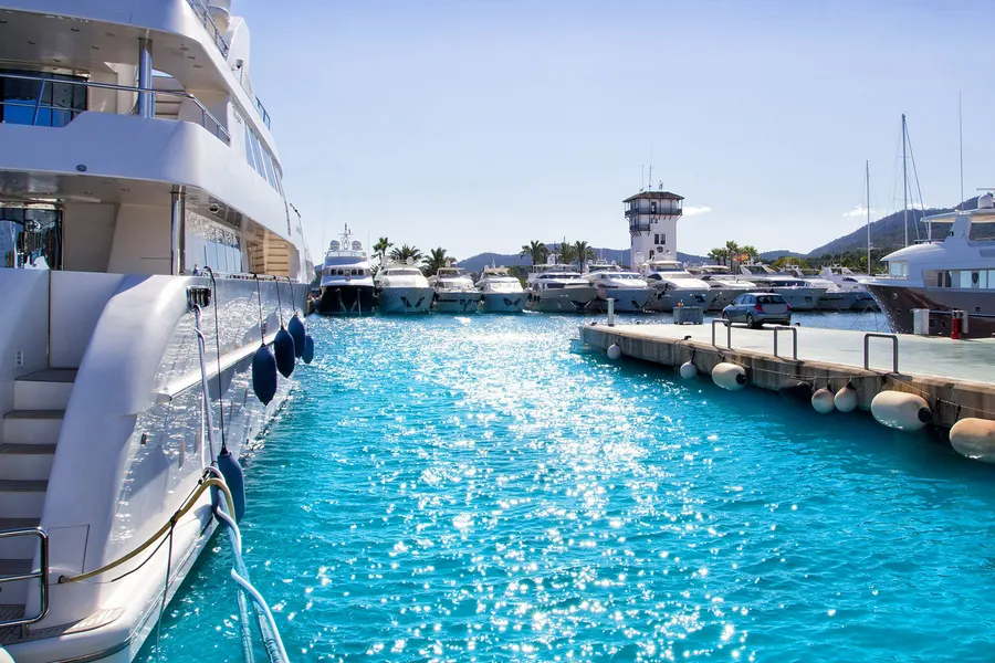 Bordwand einer sehr großen weißen Motoryacht in Puerto Portals, daneben das türkisfarbene Hafenbecken und weitere Boote am Steg.