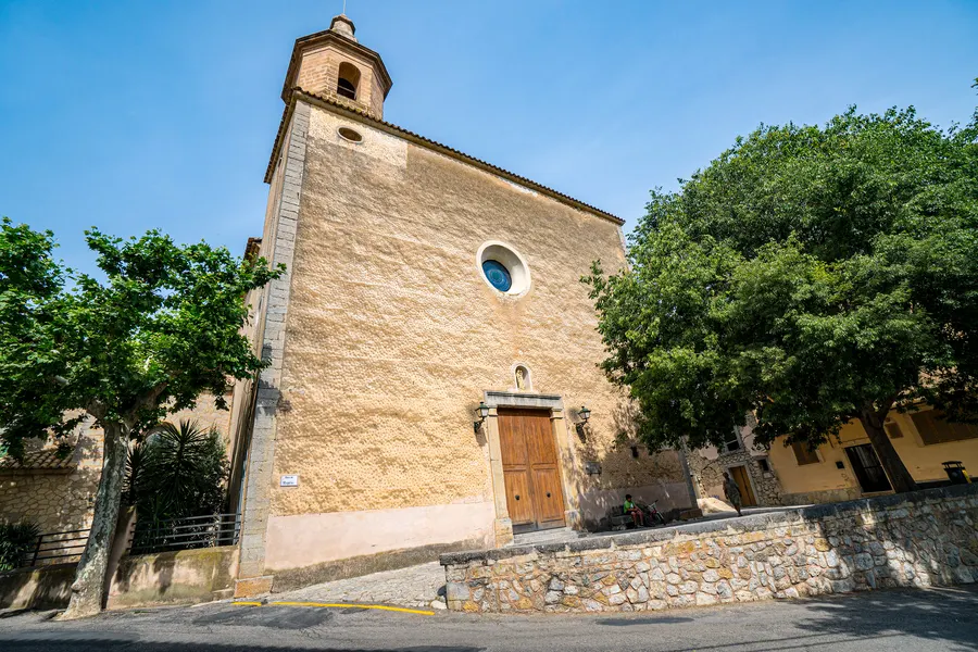Die schlichte Kirche von Puigpunyent aus hellem Stein mit rundem Fenster und Glockengiebel, davor ein Baum und eine Mauer.