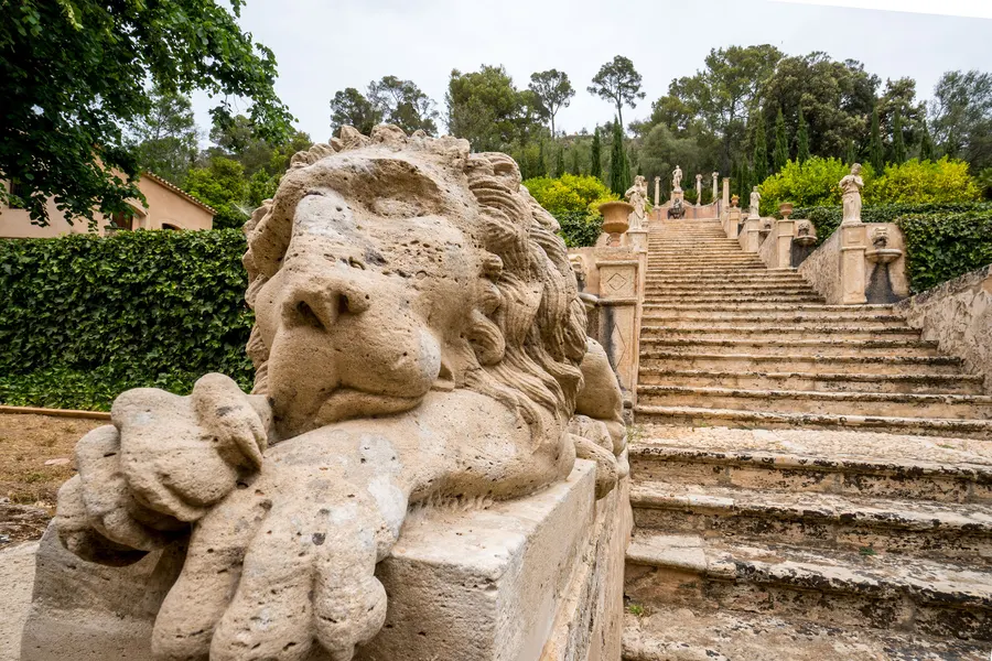 Steinerner Löwe auf der Balustrade der großen Freitreppe von Raixa, dahinter die ansteigenden Stufen zwischen Hecken und Bäumen.