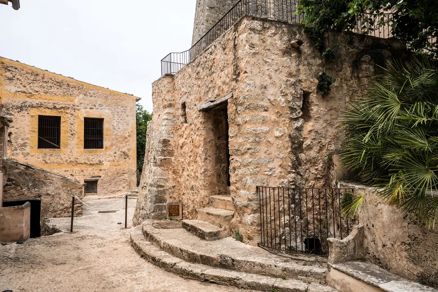 Alte Wirtschaftsgebäude aus grobem Naturstein auf dem Gut Raixa mit Steintreppe, vergitterten Fenstern und einem schmiedeeisernen Tor.
