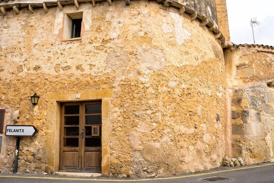 Sandsteinhaus mit gerundeter Ecke und hölzerner Tür in S'Alqueria Blanca, daneben ein Wegweiser nach Felanitx.