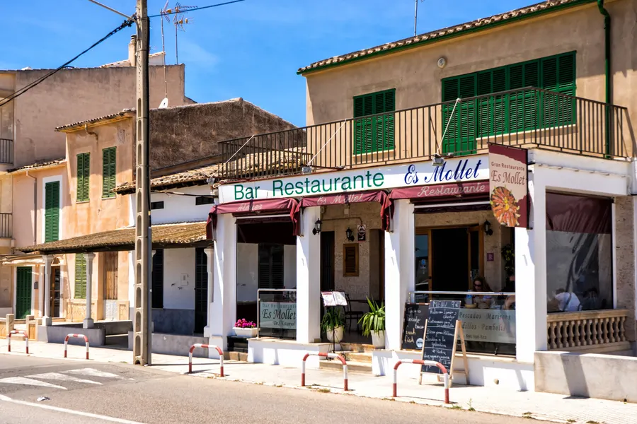 Zweigeschossiges Gebäude mit der Aufschrift Bar Restaurante in S'Estanyol de Migjorn, mit Säulen, Markisen und Tafeln davor.