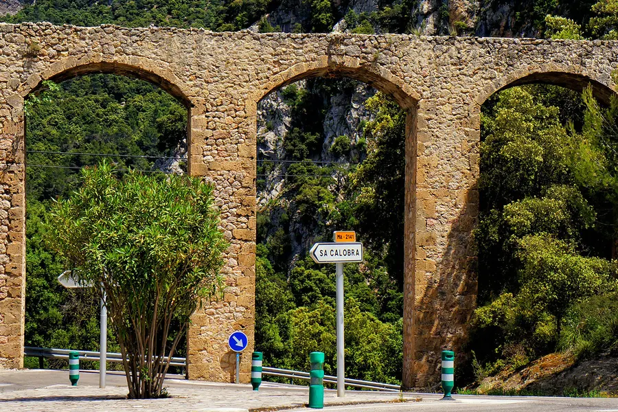Dreibogiges Natursteinviadukt an der Straße nach Sa Calobra mit einem Wegweiser davor, dahinter bewaldeter Hang.