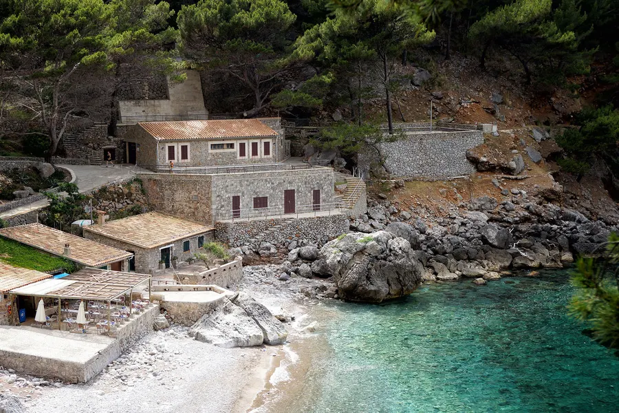 Steinerne Gebäude über der kleinen Bucht von Sa Calobra mit Terrassen, Treppen und klarem türkisfarbenem Wasser davor.
