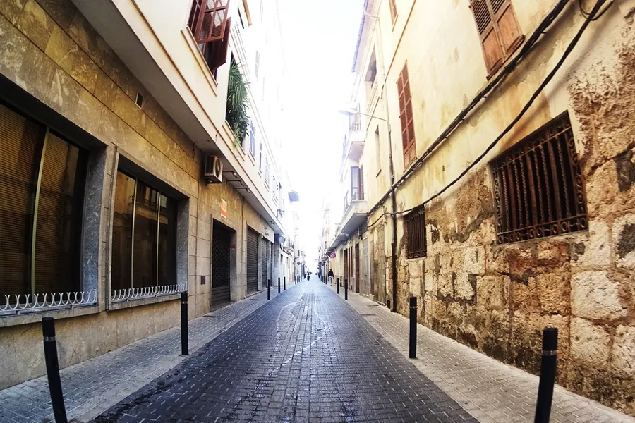 Gepflasterte Gasse in sa Pobla mit Pollern, Ladenfronten links und einer alten Bruchsteinfassade mit vergitterten Fenstern rechts.
