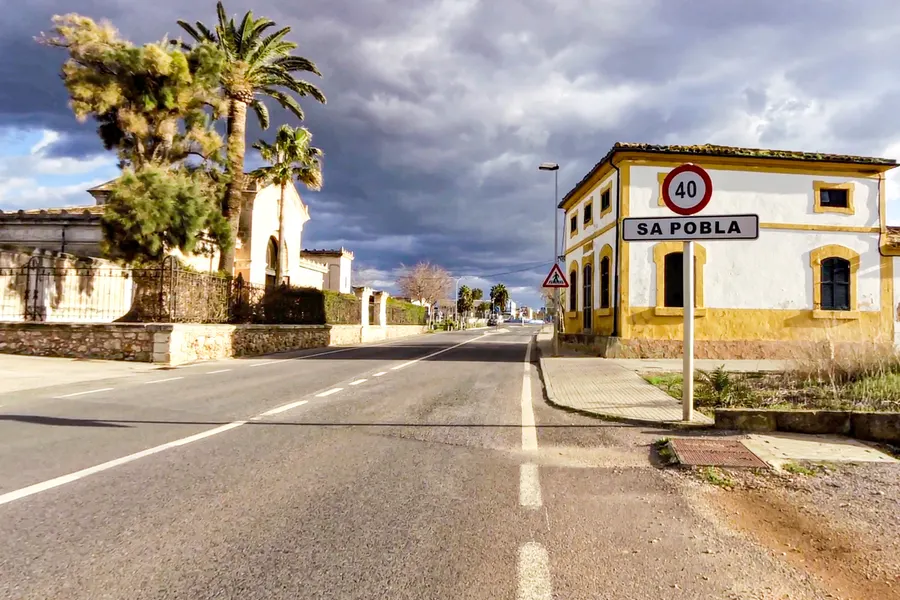 Zufahrtsstraße nach sa Pobla mit dem Ortsschild, einem gelben Eckhaus, Palmen und dunklen Wolken über der Ebene.
