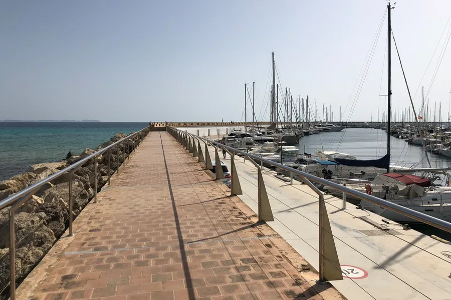 Lange gepflasterte Mole im Hafen von Sa Ràpita mit Geländer, rechts die Stege mit Segelbooten, links die Steinschüttung.
