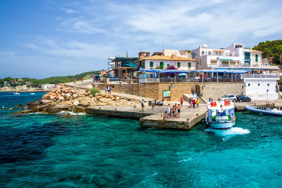 Betonanleger in Sant Elm mit einem ablegenden Fährboot, Wartenden am Kai und den Häusern des Orts auf dem Felsen dahinter.