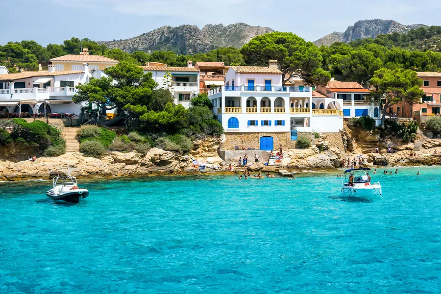 Weiße und ockerfarbene Häuser mit blauen Läden über der Felsküste von Sant Elm, davor klares türkisfarbenes Wasser mit Booten.