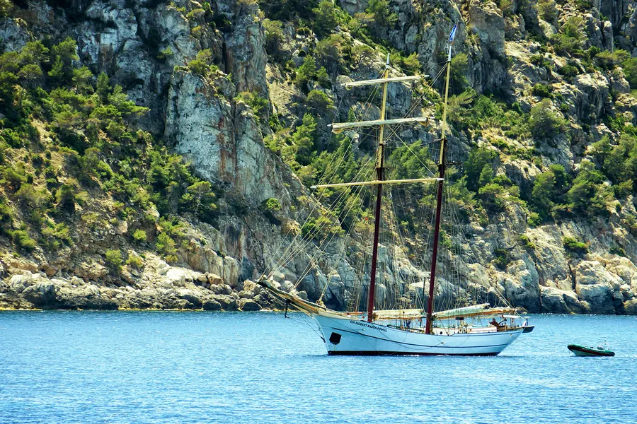 Zweimastiger Segler vor der bewaldeten Steilküste der Dragonera, im blauen Wasser vor Anker.
