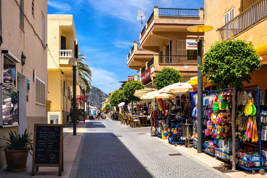 Straße in Sant Elm mit Souvenirläden, Sonnenschirmen und Kübelpflanzen, dahinter Wohnhäuser mit Balkonen.
