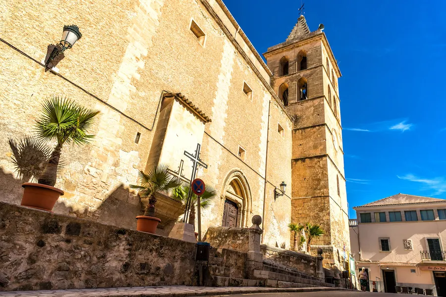 Die Pfarrkirche von Sant Joan mit hohem Glockenturm aus hellem Stein, davor eine Mauer mit Kübelpalmen und einem Kreuz.
