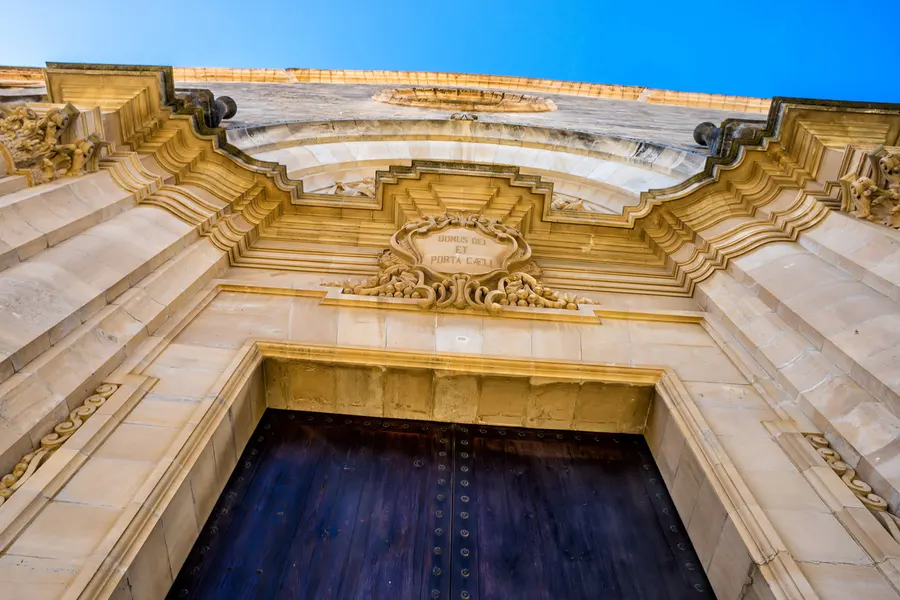 Reich profiliertes Steinportal in Sant Joan mit geschwungenem Aufsatz, Wappenkartusche und schwerer dunkler Holztür.