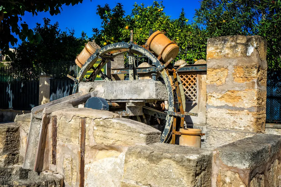 Alte Schöpfvorrichtung mit eisernem Rad und Tonkrügen über einem Steinbrunnen in einem Garten, umgeben von Zitrusbäumen.
