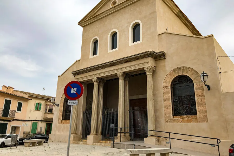 Sandfarbene Kirchenfassade in Sant Jordi mit vier Säulen vor dem Eingang, Dreiecksgiebel und drei hohen Rundbogenfenstern.