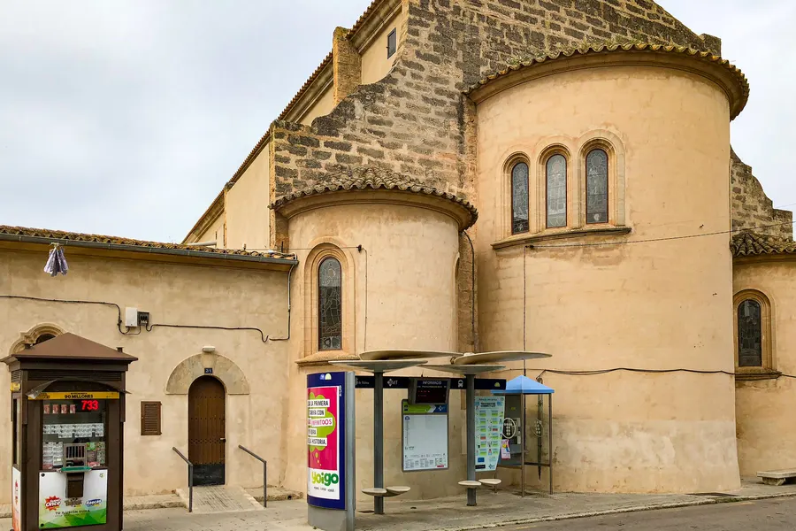 Die runde Apsis der Kirche von Sant Jordi mit Bogenfenstern, davor eine Bushaltestelle mit Fahrplan und ein Lottokiosk.

