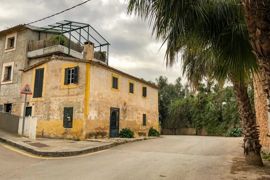 Ockergelbes älteres Wohnhaus mit dunklen Fensterläden an einer Straßenkurve in Sant Jordi, daneben Palmen und ein Verkehrsschild.
