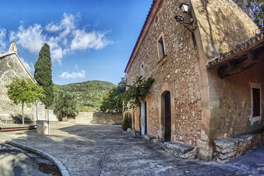 Seitenansicht von Sant Miquel mit offener Vorhalle unter hölzernem Vordach, davor der Vorplatz und eine Trockenmauer.