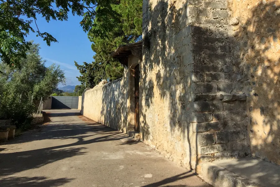 Schmale Zufahrt entlang der hohen Bruchsteinmauer von Sant Miquel, darüber Bäume und blauer Himmel.
