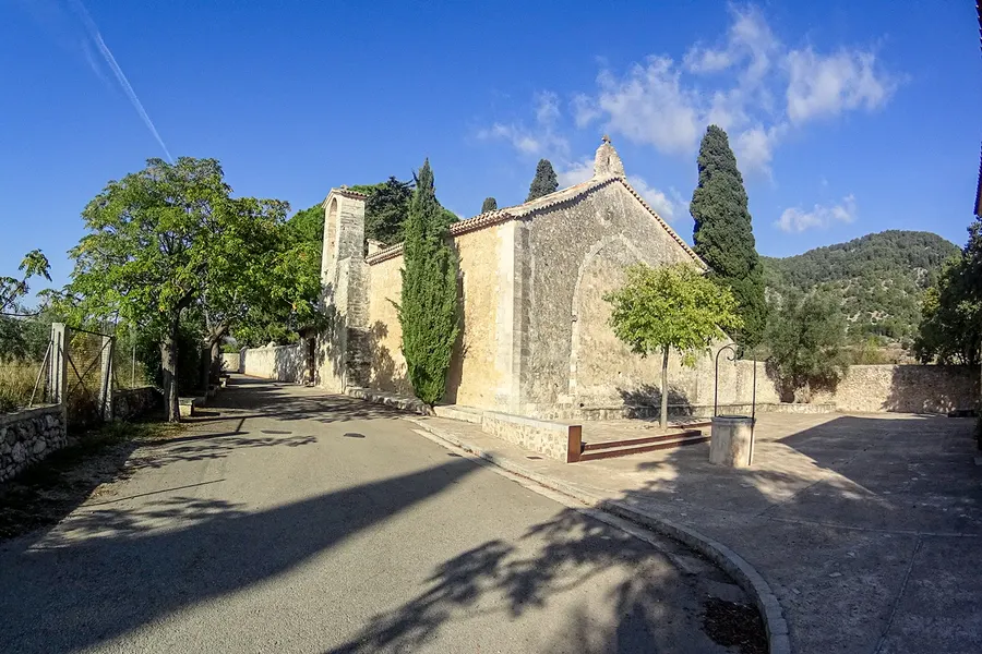 Blick von der Landstraße auf Sant Miquel de Campanet zwischen Bäumen und einer niedrigen Trockenmauer, dahinter Felder.