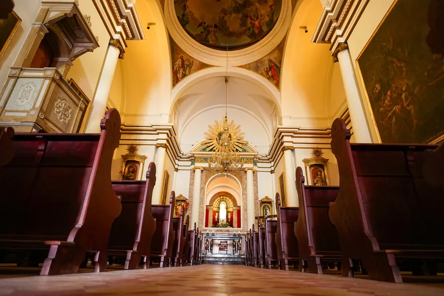 Innenraum der Wallfahrtskirche Sant Salvador mit Tonnengewölbe, Deckenfresken, dunklen Holzbänken und dem hell beleuchteten Altarraum am Ende des Mittelgangs.
