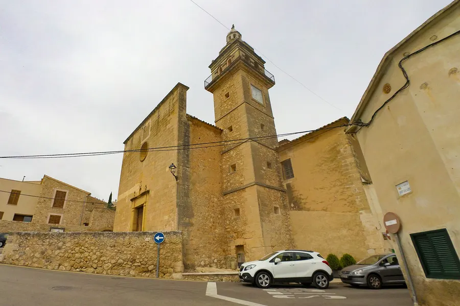 Die Kirche von Santa Eugènia mit schlankem Glockenturm und ockerfarbener Fassade, daneben ein Wohnhaus und parkende Autos.