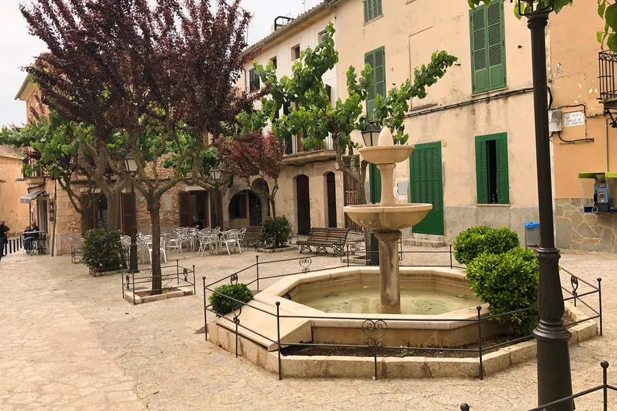 Schattiger Platz in Santa Eugènia mit rundem Brunnen, Bäumen und Natursteinhäusern mit grünen Fensterläden ringsum.