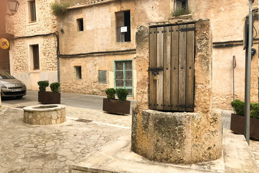 Alter steinerner Brunnen mit Ziehvorrichtung mitten in einer Gasse von Santa Eugènia, daneben parkende Autos und Natursteinhäuser.
