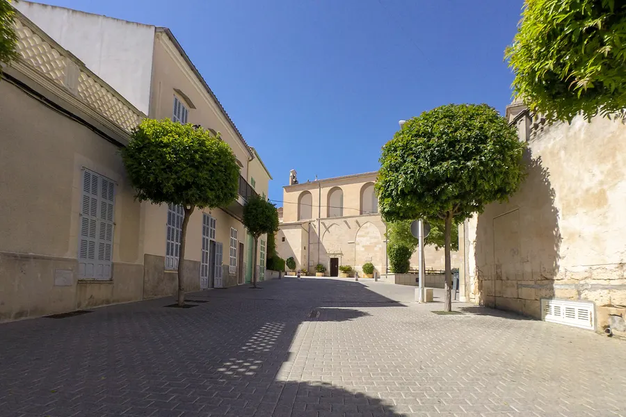 Ansteigende Gasse am Kirchplatz von Santa Margalida mit hellen Häusern, Bäumen und der Kirchenmauer auf der rechten Seite.