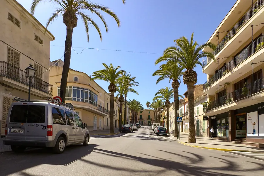 Breite Straße in Santa Margalida mit Palmenreihe, parkenden Autos und Wohnhäusern mit Rollläden.
