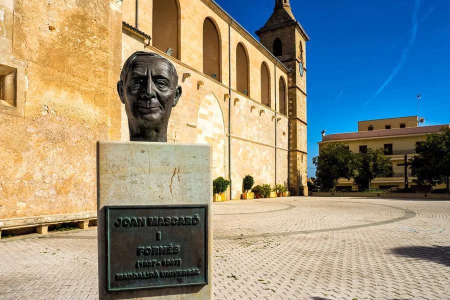 Bronzebüste auf steinernem Sockel mit Inschriftentafel an einem Platz in Santa Margalida, dahinter Häuser und blauer Himmel.
