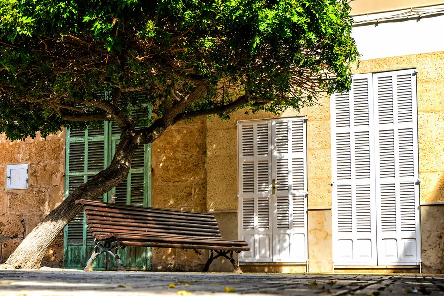 Sandsteinfassade mit grünen Fensterläden in Santa Margalida, überwachsen von Weinlaub, davor eine Bank im Schatten.
