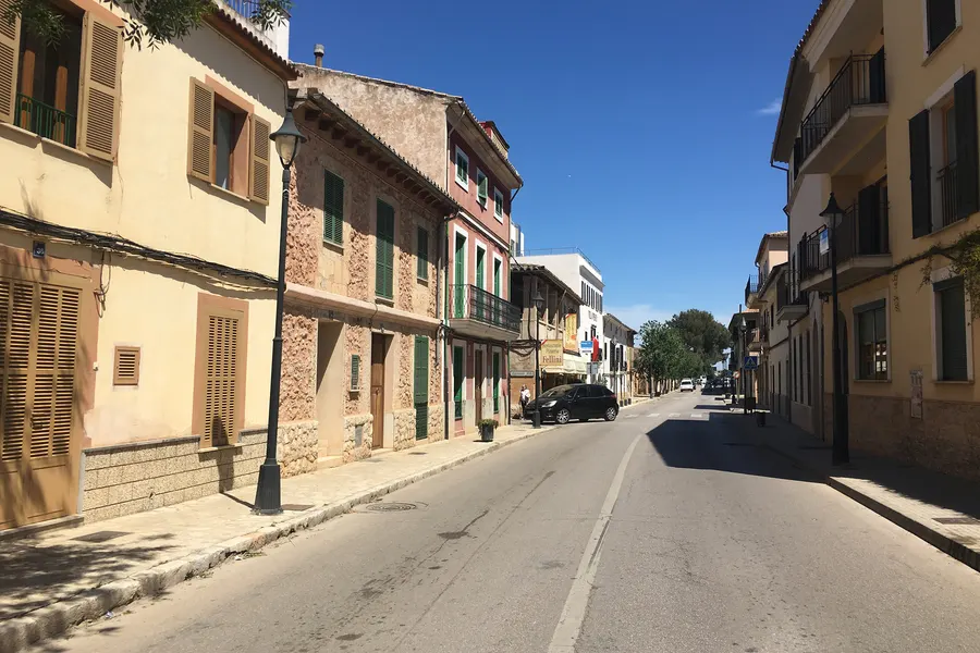 Häuserzeile in Santa Maria del Camí mit Balkonen, grünen Fensterläden und Läden im Erdgeschoss entlang der Hauptstraße.
