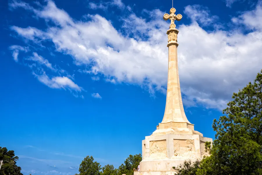 Hohes steinernes Denkmal in Form einer schlanken Säule mit vergoldetem Kreuz auf der Spitze, vor blauem Himmel mit Wolken.