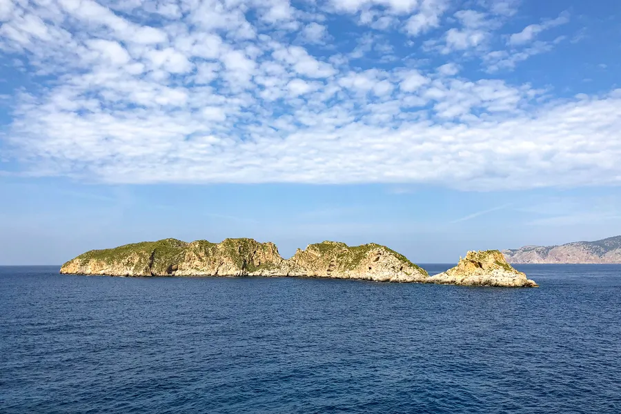 Die flachen bewachsenen Felsinseln der Illes Malgrats vor der Küste, vom Wasser aus gesehen, unter hohem Wolkenhimmel.