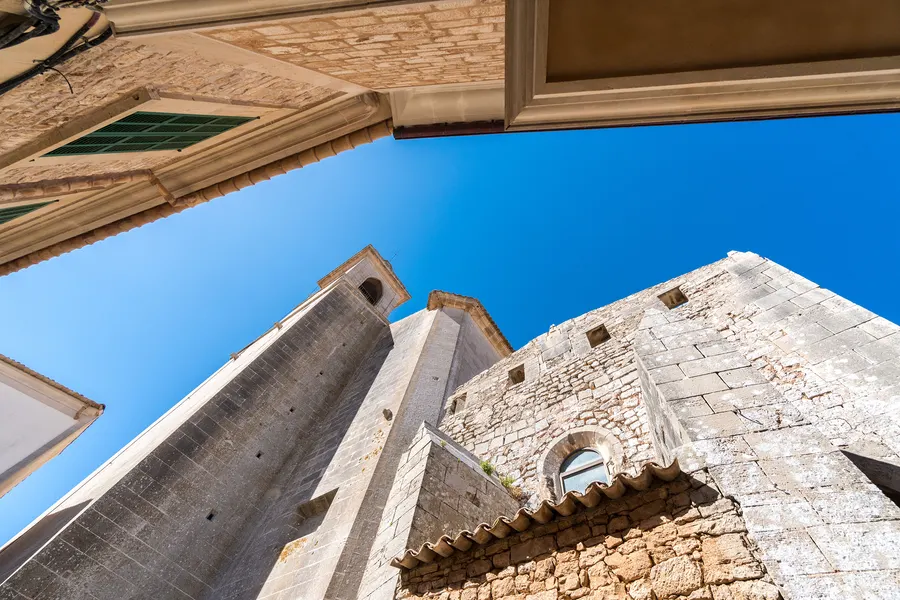 Der massive Glockenturm von Sant Andreu in Santanyí steil von unten fotografiert, daneben das Kirchenschiff mit Rundfenster.
