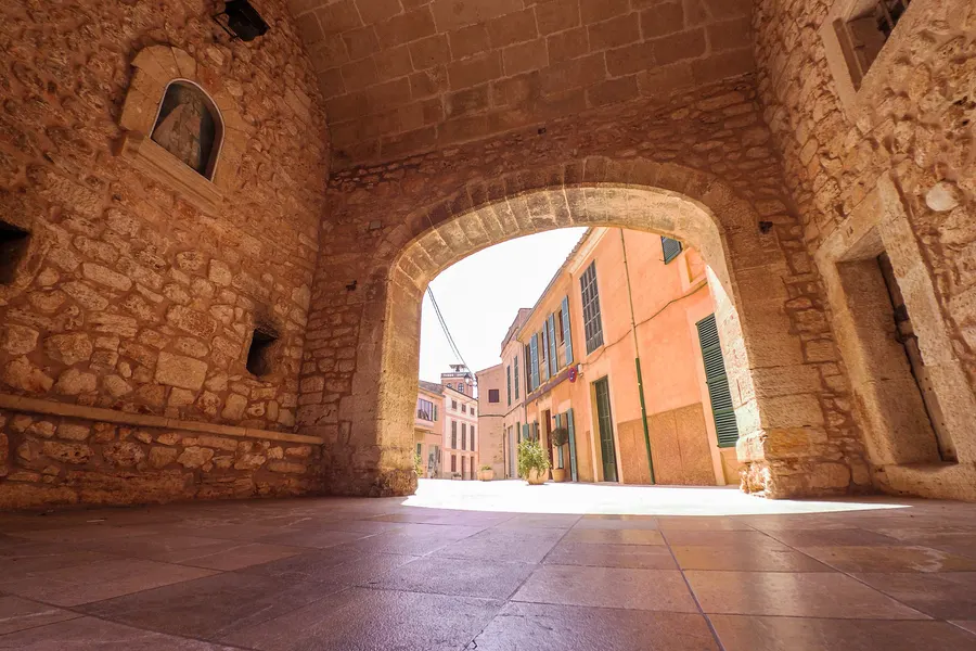 Blick aus dem Schatten des Torbogens der Porta Murada hinaus auf die sonnenbeschienene Gasse mit ockerfarbenen und rosa Häusern.