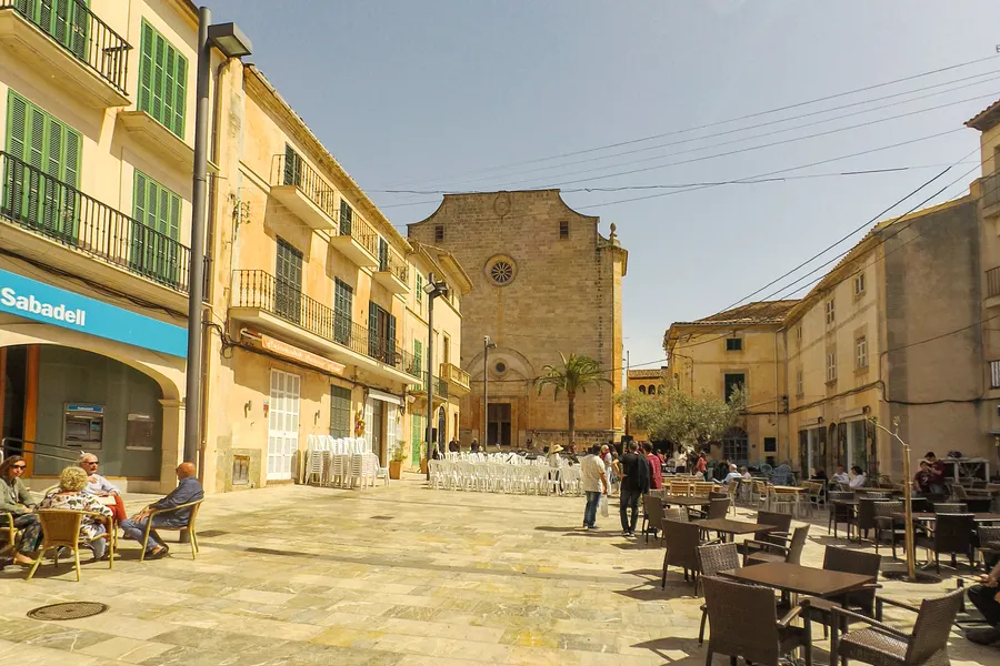 Weiter Platz in Santanyí mit Café-Terrassen und Gästen, dahinter die wuchtige Sandsteinfassade der Kirche Sant Andreu.