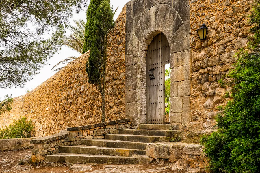 Steinerne Treppe hinauf zu einem spitzbogigen Tor in der hohen Klostermauer des Santuari de la Consolació, ringsum Bewuchs.