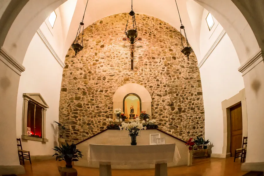 Innenraum der Kapelle mit Tonnengewölbe aus hellem Naturstein, weiß getünchtem Altarraum, Blumenschmuck und schlichten Holzstühlen.
