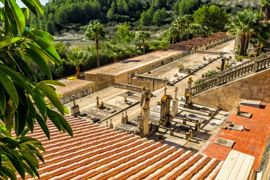 Blick über ein Ziegeldach hinunter auf den terrassierten Friedhof von Sant Joan mit Grabplatten, Kreuzen und Wandnischen, dahinter Palmen und Felder.
